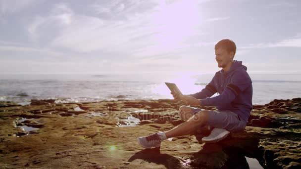Jonge man heeft een video-chat met zijn vriend via zijn tablet pc zittend op een strand — Stockvideo