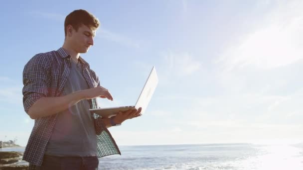 Giovane bello è in chat con qualcuno utilizzando il suo computer portatile mentre in piedi su una spiaggia e guardando bellissimo paesaggio — Video Stock