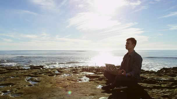 Jovem atraente está meditando enquanto está sentado em uma praia com seu laptop — Vídeo de Stock