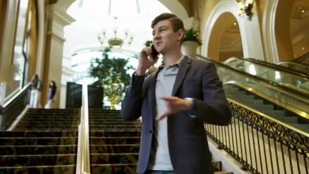 Молодой человек разговаривает по мобильному телефону в помещении рядом с лестницей — стоковое видео