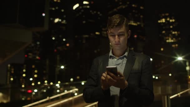 Bonito jovem estudante está usando seu smartphone para redes sociais enquanto está de pé em uma rua — Vídeo de Stock