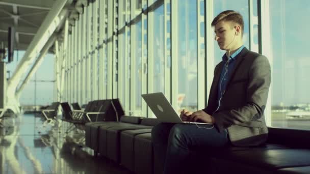 Young man is sitting in airport with a laptop and an earphones — Stock Video