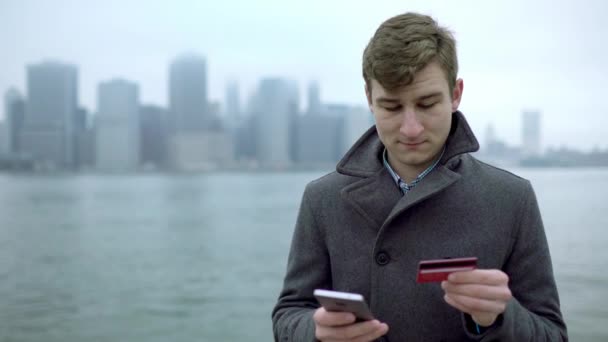 Привлекательный молодой человек покупает что-то в интернете, стоя возле реки Гудзон с Манхэттенским островом за спиной. — стоковое видео