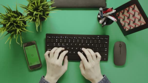 Чоловік, який друкує на клавіатурі, сидячи за столом з клавішею Chrome і використовуючи свій смартфон з зеленим екраном, вид зверху вниз — стокове відео