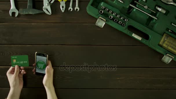 Een man zit aan een tafel naast hem is een koffer met de hulpmiddelen en hij wordt steeds om iets op het internet te kopen met behulp van een bankkaart. — Stockvideo