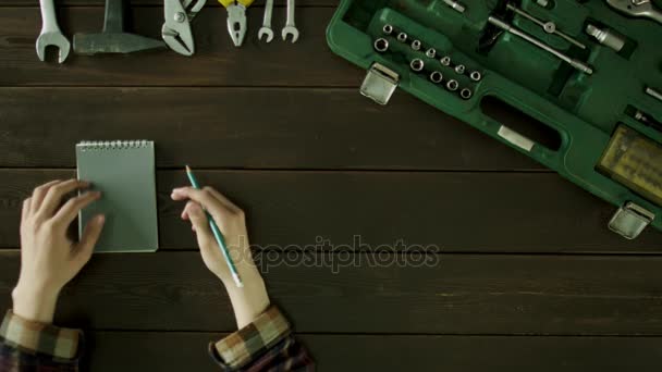 Ένας άνδρας κάθεται σε ένα τραπέζι κοντά τα εργαλεία είναι και σκέφτεται ότι για να γράψετε σε ένα σημειωματάριο. — Αρχείο Βίντεο