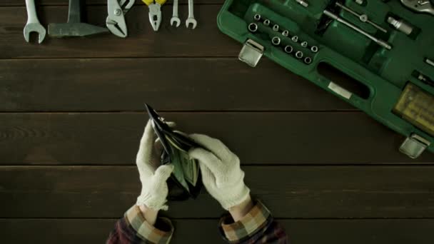 Un hombre se sienta en una mesa cerca de las herramientas son y saca dinero de su cartera, sus manos en guantes . — Vídeos de Stock
