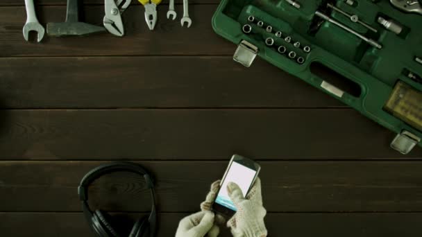 Ένας άνδρας κάθεται σε ένα τραπέζι κοντά τα εργαλεία και παίζουν μουσική στο τηλέφωνό σας μέσω των ακουστικών. — Αρχείο Βίντεο