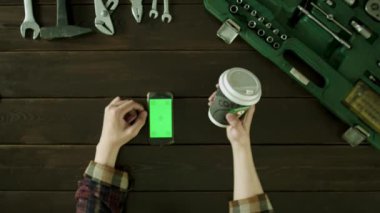 Bir adam araç kutusu yakınındaki bir masada oturur ve kahve içer ve telefonda bir şey izlemek.