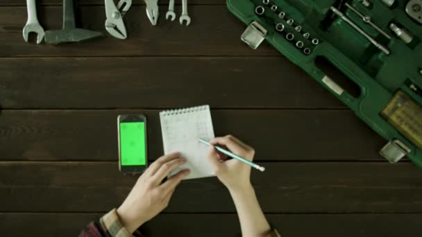男はツールボックスの近くのテーブルに座っているし、携帯電話からノートに何かを書いて. — ストック動画
