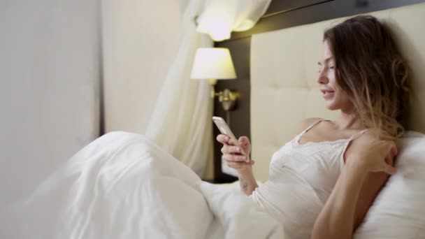 Joven hermosa mujer acostada en la cama y comprobando las redes sociales en un teléfono inteligente — Vídeo de stock