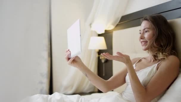 Женщина ведет видео-разговор через Интернет через свой планшетный компьютер, пока лежит на кровати — стоковое видео