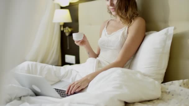Yatakta kahve yudumlarken dizüstü bilgisayar kullanan genç güzel kadın — Stok video