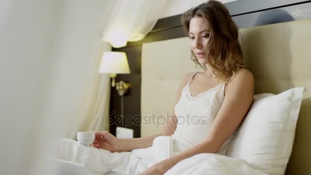 Linda mulher lendo notícias em seu laptop enquanto bebe café na cama — Vídeo de Stock