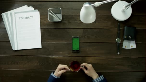 Pohled shora. Člověk pije čaj u stolu office a používá svůj telefon. — Stock video