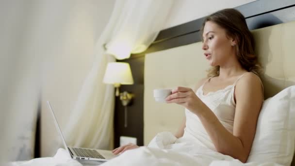 Ung kvinde gør video oversættelse på sin bærbare computer, mens du drikker kaffe i sengen – Stock-video