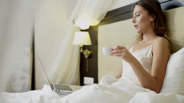 Prachtige vrouw doen video vertaling op haar laptop terwijl het drinken van koffie in bed — Stockvideo