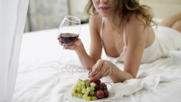 Femme boit du vin rouge et mange du raisin tout en étant couchée au lit — Video