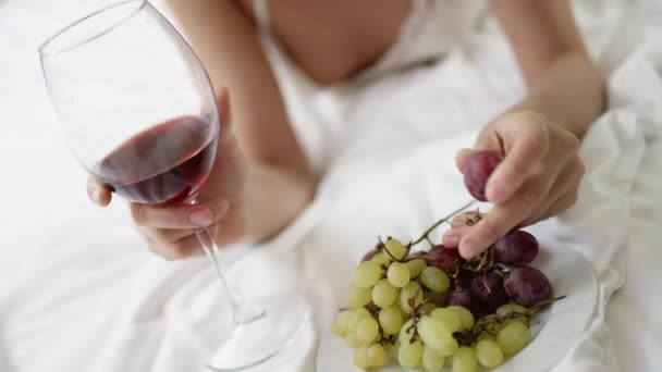 Woman drinks wine and eats grape while lying on bed — Stock Video