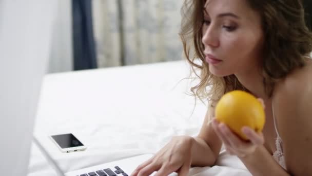 Vrouw met behulp van haar laptop en een oranje op bed holding — Stockvideo