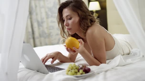 Mujer joven leyendo noticias en su portátil y sosteniendo y naranja con uvas en el plato cerca de ella — Vídeo de stock