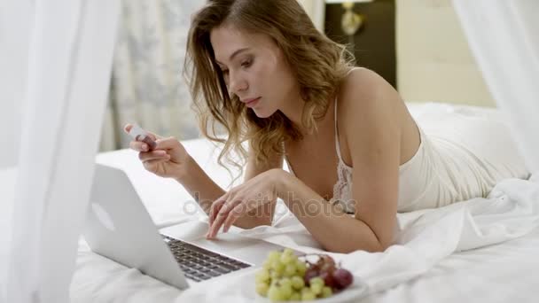 Woman shopping online while lying on her bed by using her laptop and credit card — Stock Video