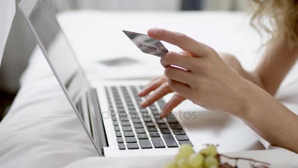 Mujer acostada en la cama y comprando algo en línea mediante el uso de tarjeta de crédito y portátil — Vídeos de Stock