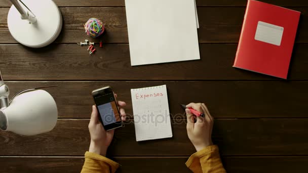 Der Mann am Schreibtisch klebt Aufkleber in das Notizbuch und benutzt sein Handy neben dem Ordner, der Lampe, dem Ball, dem Büro und den Knöpfen. — Stockvideo