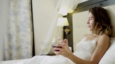 Bir bardak kırmızı şarap ve onun smartphone haber okuma ile yatakta kadın yatıyor