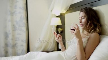 Bir bardak kırmızı şarap ve sosyal ağ onun smartphone üzerinde kontrol ile yatakta beyaz pijama giymiş kadın yatıyor