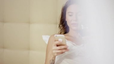 Güzel kadın smartphone ile yatakta yatıyor ve içecekler cam dışında kırmızı şarap