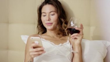 Güzel kadının yatak ve sosyal ağ bir smartphone cep telefonu ile kontrol üzerinde yatıyor ve içecekler cam dışında kırmızı şarap