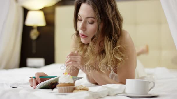 Preciosa mujer escribiendo algo en su cuaderno con una taza de café y magdalena en la cama — Vídeos de Stock