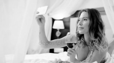 Çekici kadın kırmızı şarap içiyor ve siyah ve beyaz yatakta kendini resmi onun smartphone için alır