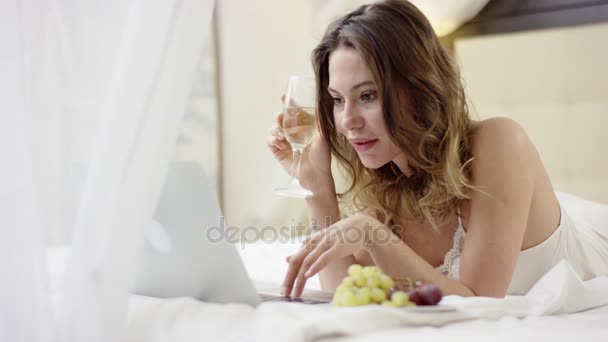 Mulher bonita deitada na cama com um copo de vinho branco e lendo notícias em seu laptop — Vídeo de Stock