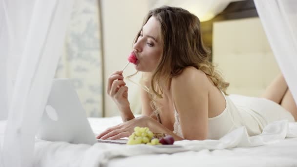 Сексуальная женщина лежит на кровати и облизывает жесткие конфеты во время просмотра фильма на ноутбуке — стоковое видео