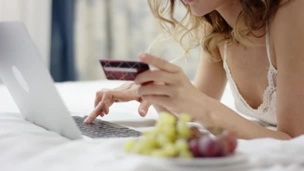 Женщина в сонной одежде лежит на кровати и использует свой ноутбук и кредитную карту для онлайн-банкинга — стоковое видео