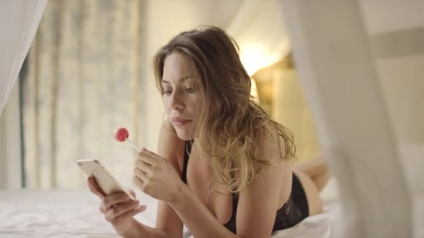 Donna affascinante utilizzando smartphone e leccare caramelle dure sul letto — Video Stock