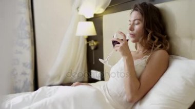 Genç kadın pijamalar yatağın üstüne kırmızı şarap içme smartphone kullanarak