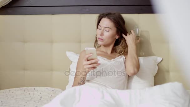 Ελκυστική γυναίκα ξαπλωμένη στο κρεβάτι με ένα ποτήρι σαμπάνια και ένα smartphone — Αρχείο Βίντεο