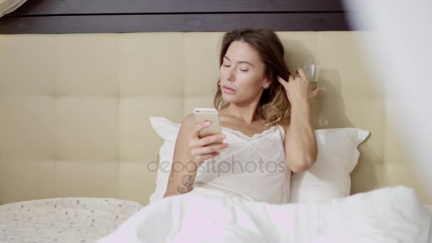 Atractiva mujer acostada en la cama con una copa de champán y un teléfono inteligente — Vídeo de stock