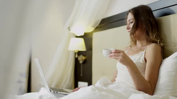 Mujer teniendo un video chat en su computadora portátil en la cama mientras bebe café — Vídeos de Stock