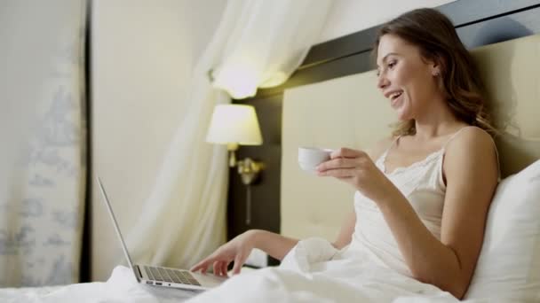 Молодая женщина делает видео-перевод на свой ноутбук на кровати во время питья кофе — стоковое видео