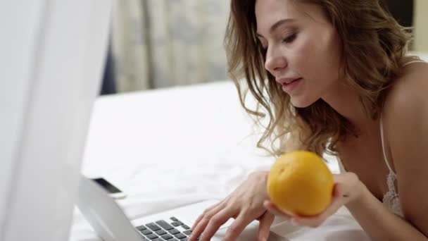 Vrouw met een laptop en oranje op bed — Stockvideo