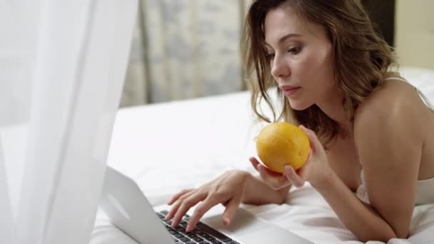 Vacker kvinna med en laptop och orange på säng — Stockvideo