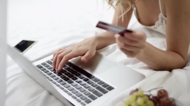 Wunderschöne Frau mit Laptop und Kreditkarte für Online-Einkäufe auf dem Bett mit Teller Trauben in ihrer Nähe — Stockvideo