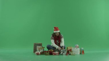 Noel hediyeleri arasında oturan ve birini chroma anahtar arka plan üzerinde çalışırken Noel şapka çekici genç kadın
