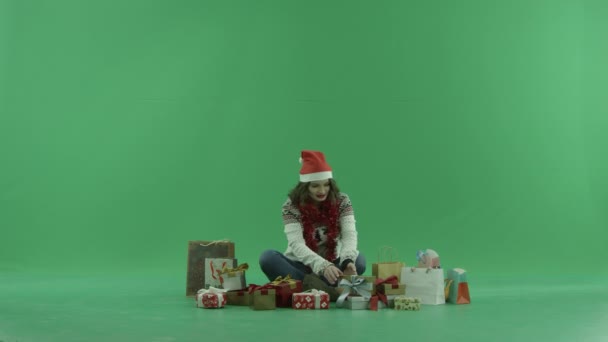 Ελκυστική νεαρή γυναίκα με καπέλο Χριστουγέννων κάθεται μεταξύ Χριστούγεννα δώρα και προσπαθεί να επιλέξει ένα, chroma κλειδί στο φόντο — Αρχείο Βίντεο