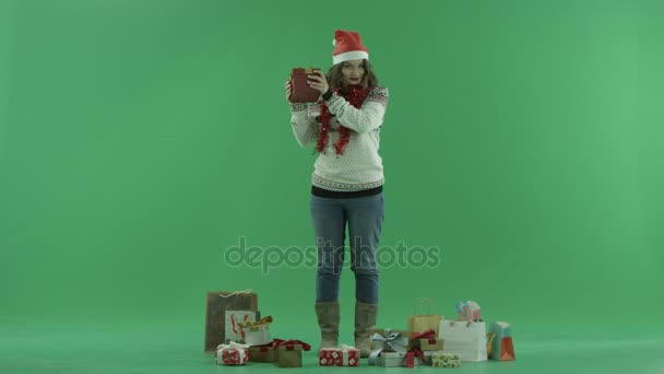 Bella giovane donna in cappello di Natale cercando di indovinare cosa si trova nella sua scatola regalo di Natale, chiave cromatica su sfondo — Video Stock