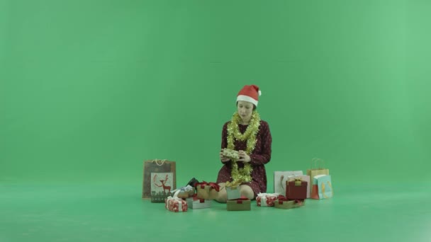 Seorang wanita muda sedang duduk dan membuka hadiah Natal di dekat dirinya di layar hijau — Stok Video